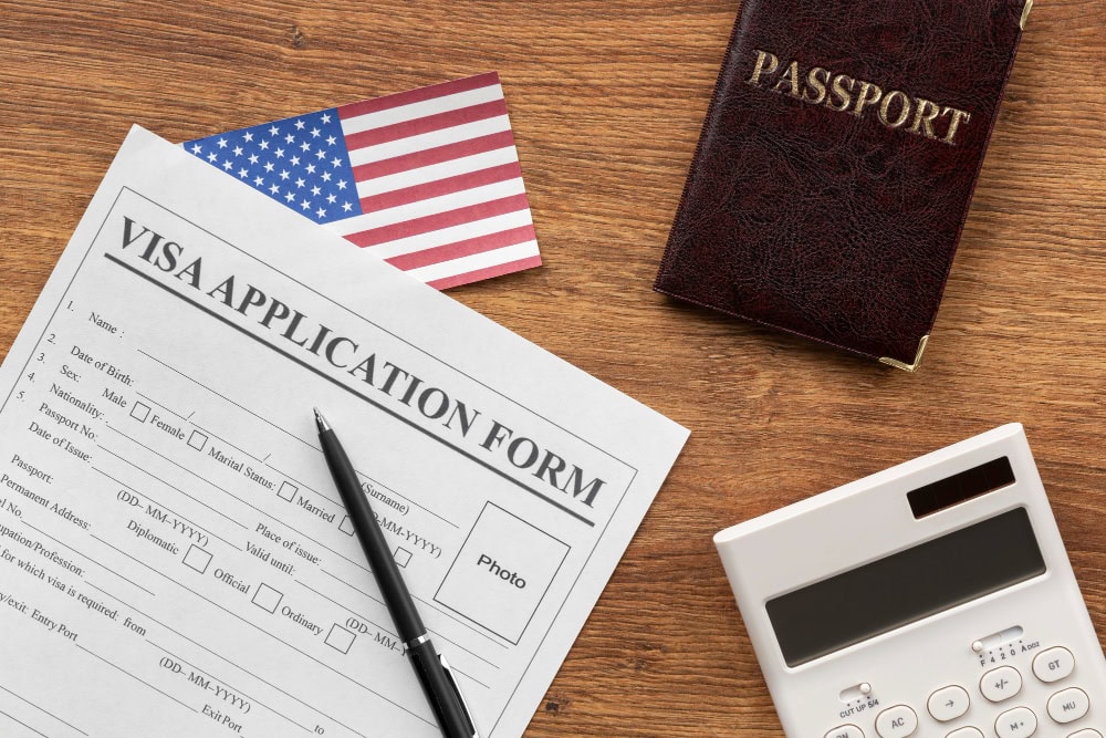 A visa application form, a passport, a calculator and pen sit on a desk next to a small American flag, symbolizing immigration law.